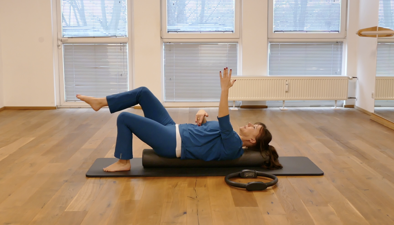Kerstin Bredehorn liegt in Rückenlage auf einer Pilates Rolle, ein Bein ist angewinkelt abgehoben, der gegenüberliegende Arm zeigt zur Decke